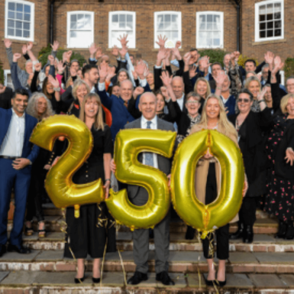 A group of people hold gold balloons forming "250" on a staircase in front of a brick building, celebrating an event. - Home Instead