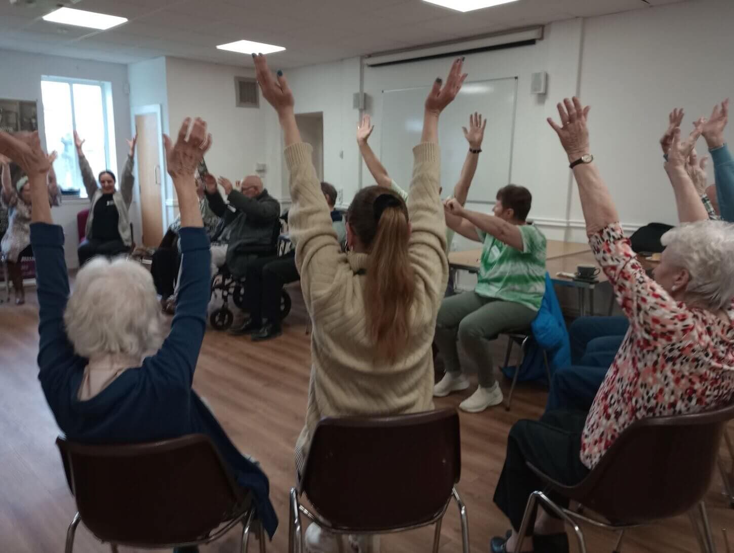 A group of elderly people in a circle, raising their arms during a seated exercise session in a bright room. - Home Instead