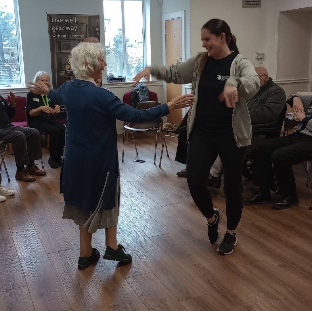 Two women, one elderly and one younger, dancing joyfully together in a community room with seated onlookers. - Home Instead
