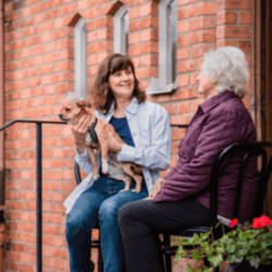 Two women sit on a porch chatting; one holds a small dog while the other listens, both appearing happy. - Home Instead
