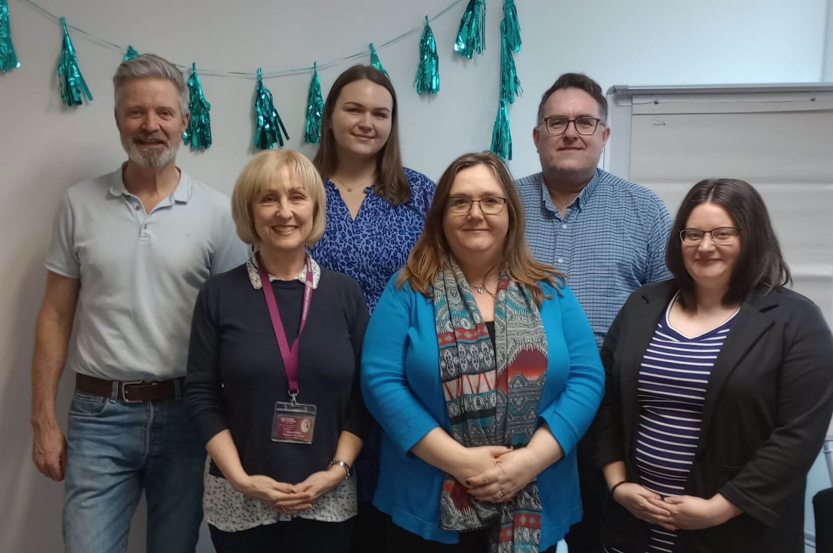 A group of six people standing indoors with teal decorations in the background. - Home Instead