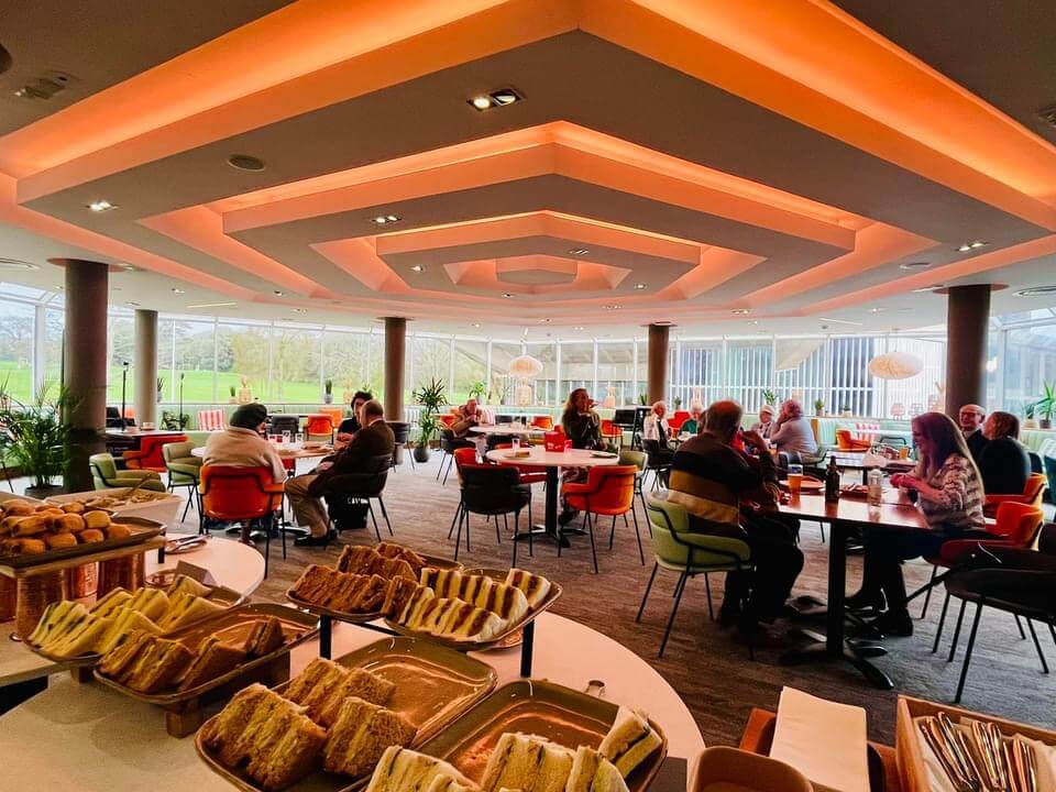 People seated at tables in a modern, well-lit cafe with sandwiches and pastries in the foreground. - Home Instead