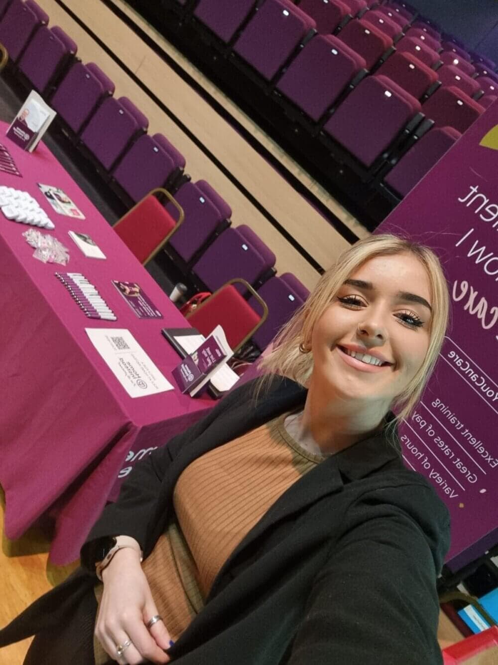 A woman smiling in front of a table with promotional materials and an empty auditorium with purple seats in the background. - Home Instead