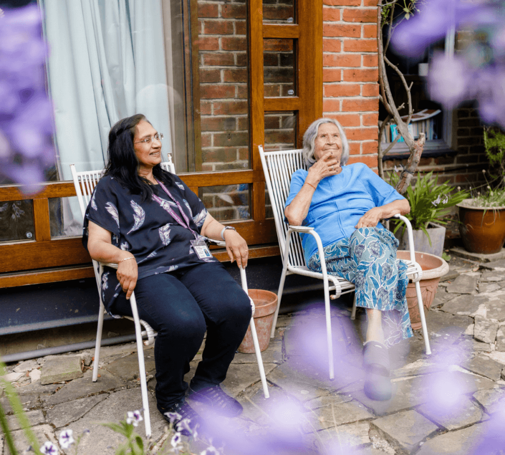 Two women sitting on chairs outside a house, surrounded by flowers, smiling and enjoying the day. - Home Instead