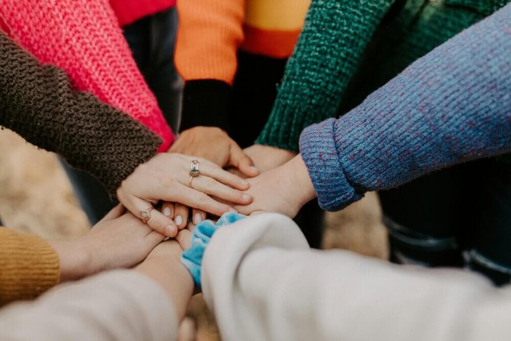 A diverse group of people placing their hands together in a circle, wearing colorful sweaters. - Home Instead