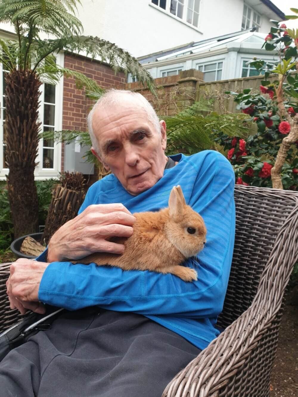 An elderly man sits in a wicker chair, holding a brown rabbit in an outdoor garden setting. - Home Instead