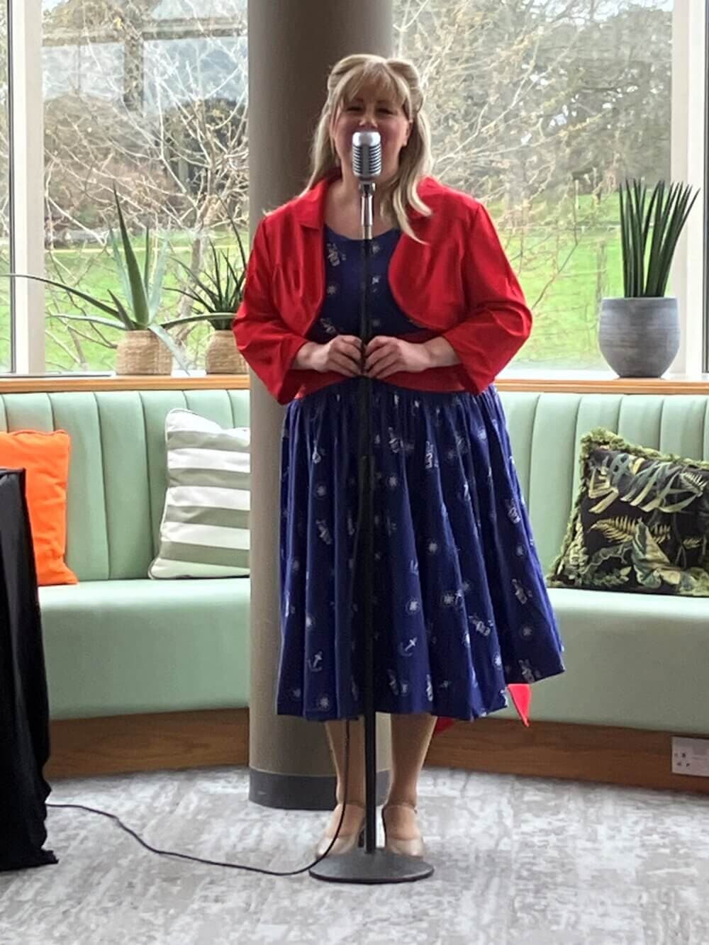 A woman in a blue dress and red jacket sings into a vintage microphone in a brightly lit room with green seating. - Home Instead
