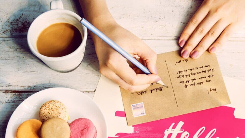 Person writing a postcard at a table with a cup of coffee and assorted macarons. - Home Instead