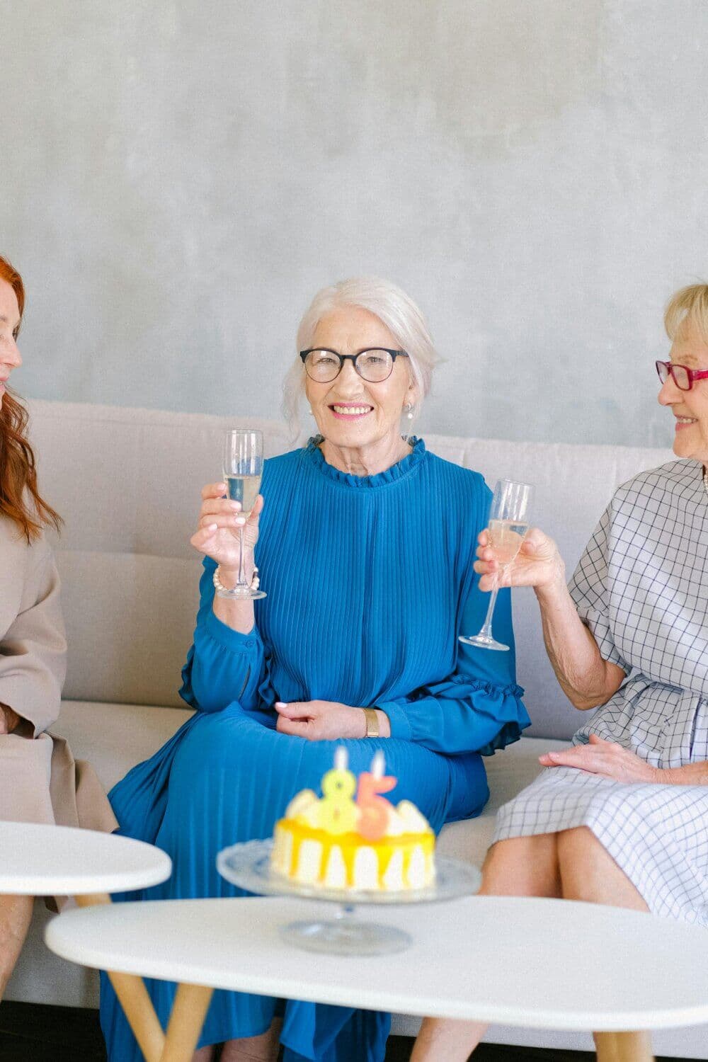 Three elderly women raising champagne glasses, with a small yellow cake in the foreground, sitting on a sofa and smiling. - Home Instead