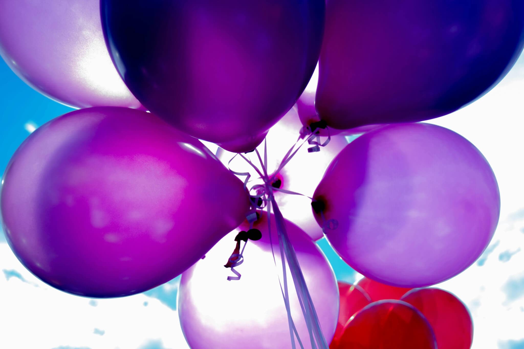 Close-up of purple balloons tied together, floating against a bright blue sky with white clouds. - Home Instead