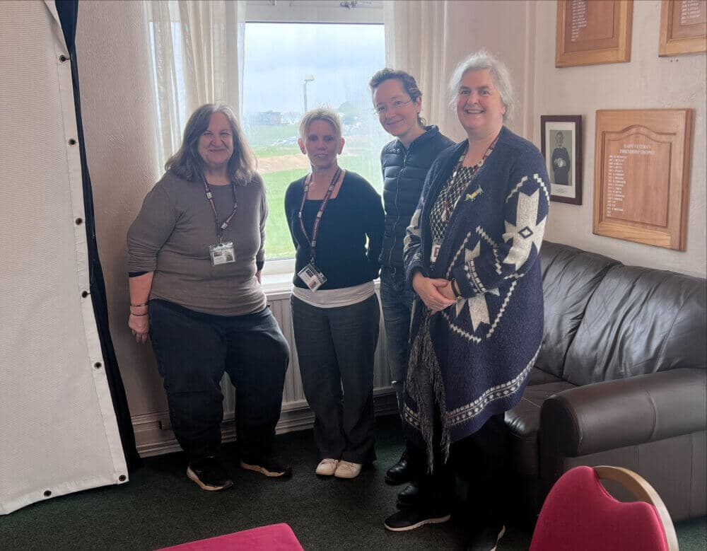 Four people stand smiling together in a cozy room with a window in the background. - Home Instead
