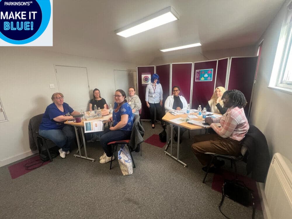 A group of people sitting around two tables in a room with red dividers, engaged in a meeting or discussion. - Home Instead