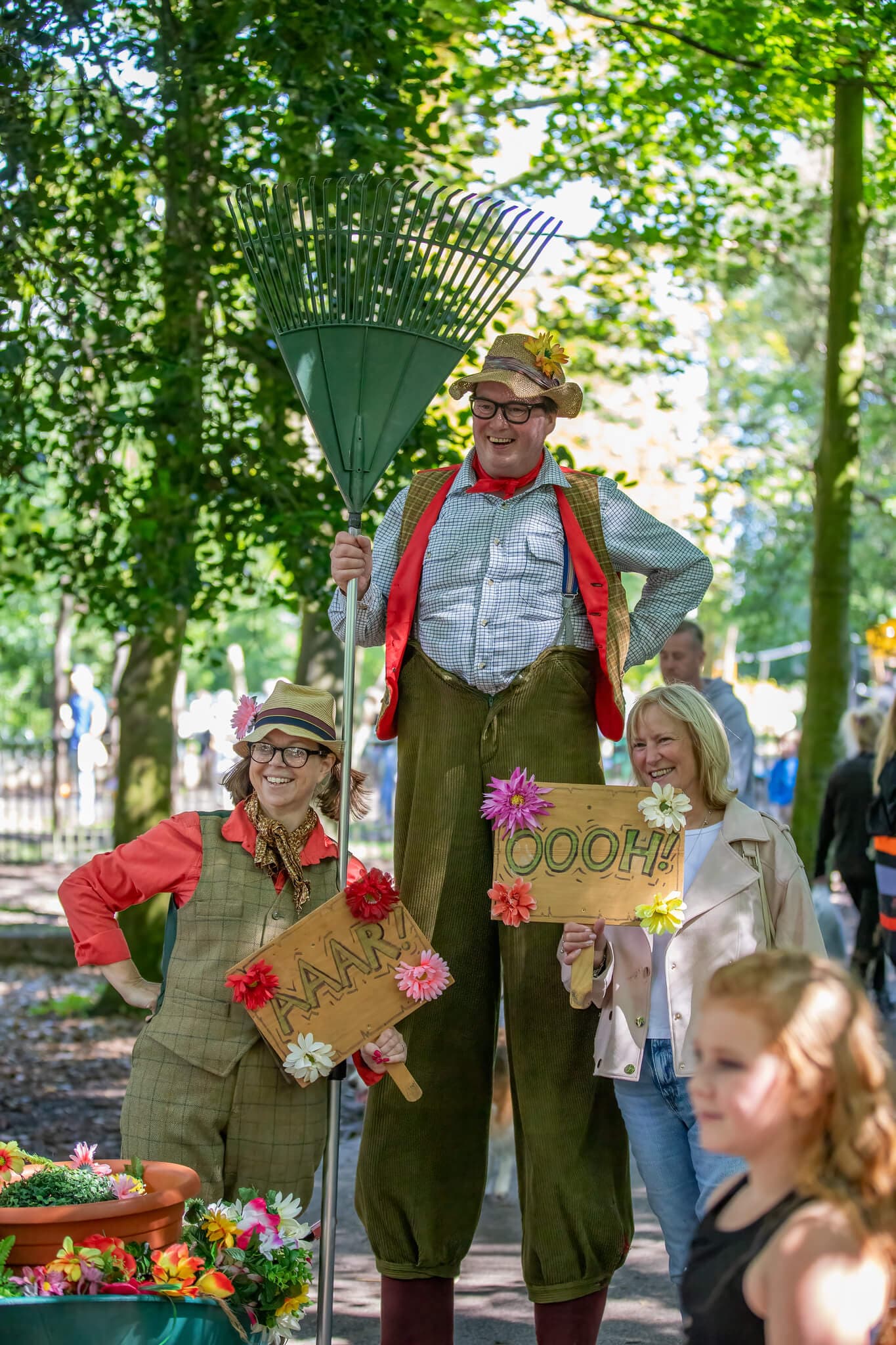 Two performers in colorful costumes, one on stilts, hold "Aah" and "Oooh" signs at an outdoor event with attendees. - Home Instead