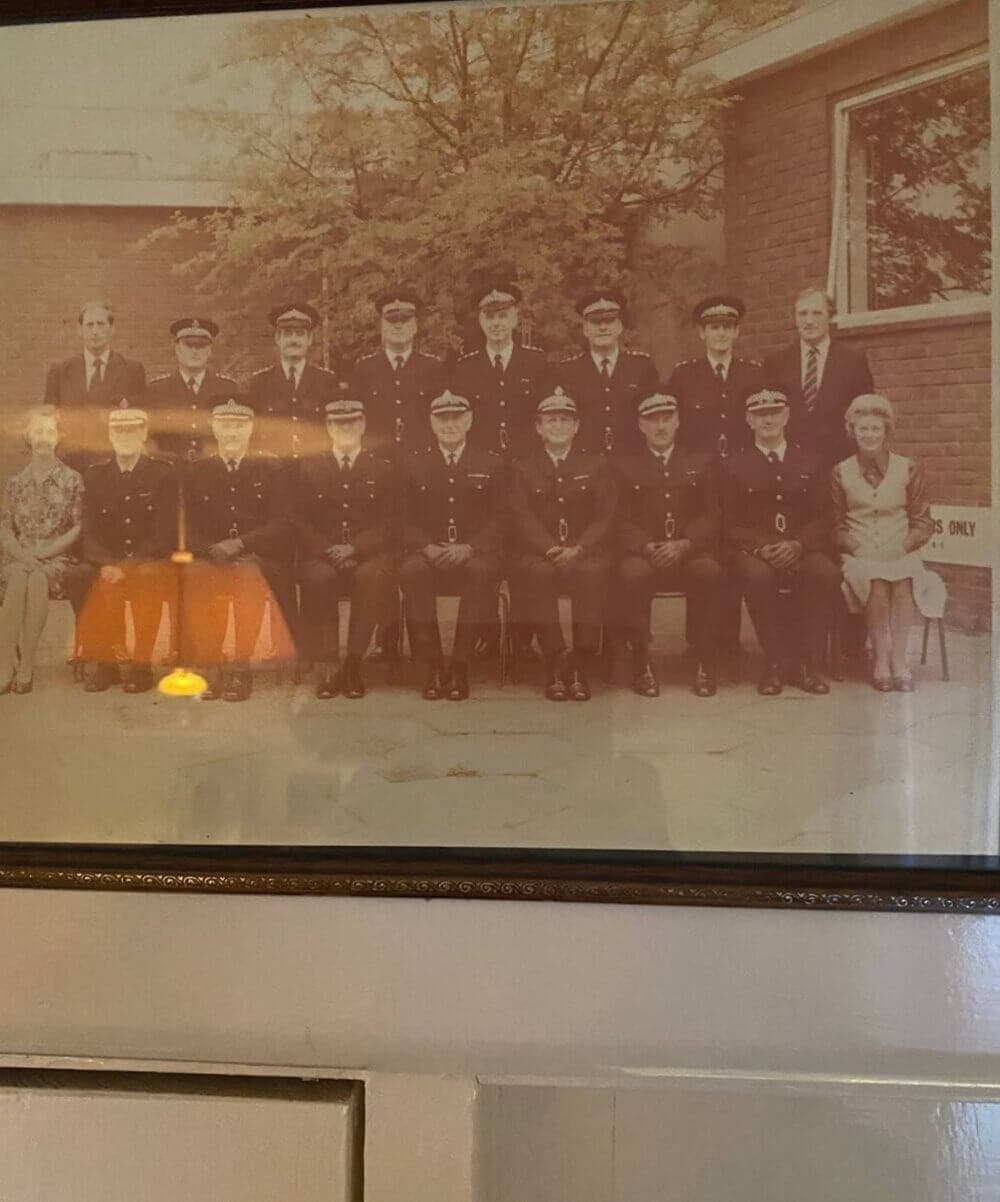 A vintage photo of 14 uniformed individuals seated and standing in front of a building, with 3 people in civilian clothes. - Home Instead