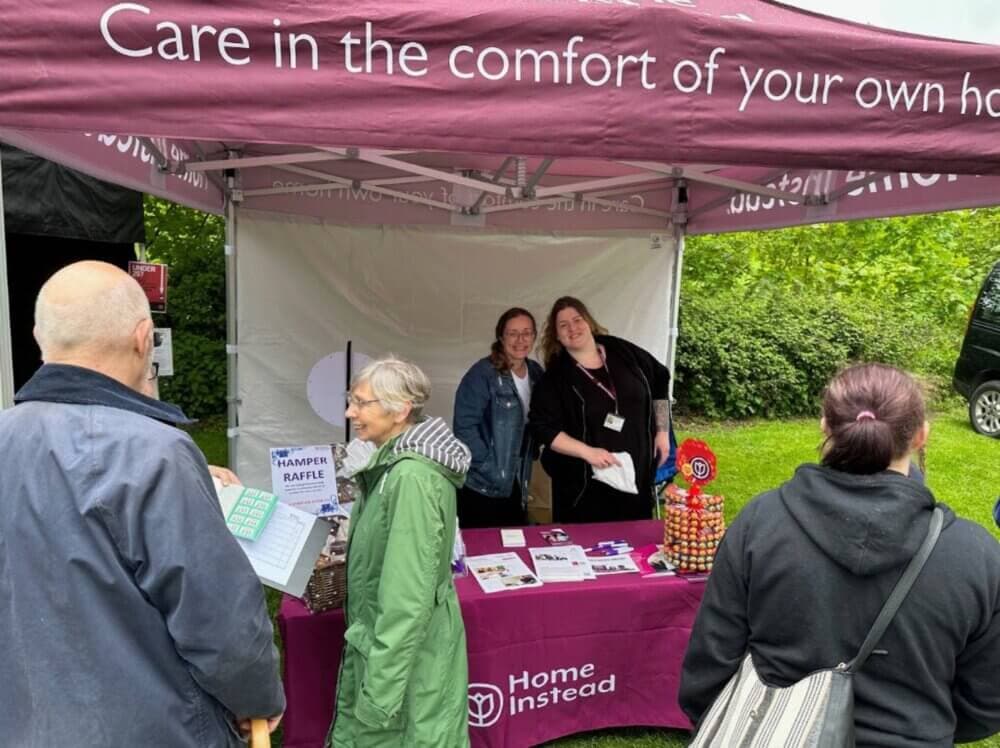 People visiting a stall with a purple canopy displaying "Home Instead," featuring brochures and a raffle sign. - Home Instead
