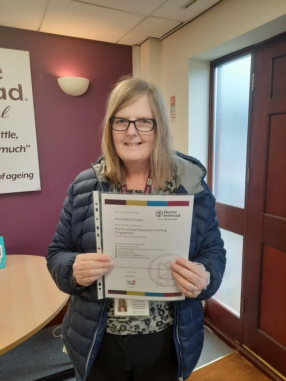 A person wearing glasses and a blue jacket holds a certificate and smiles while standing in an indoor setting. - Home Instead