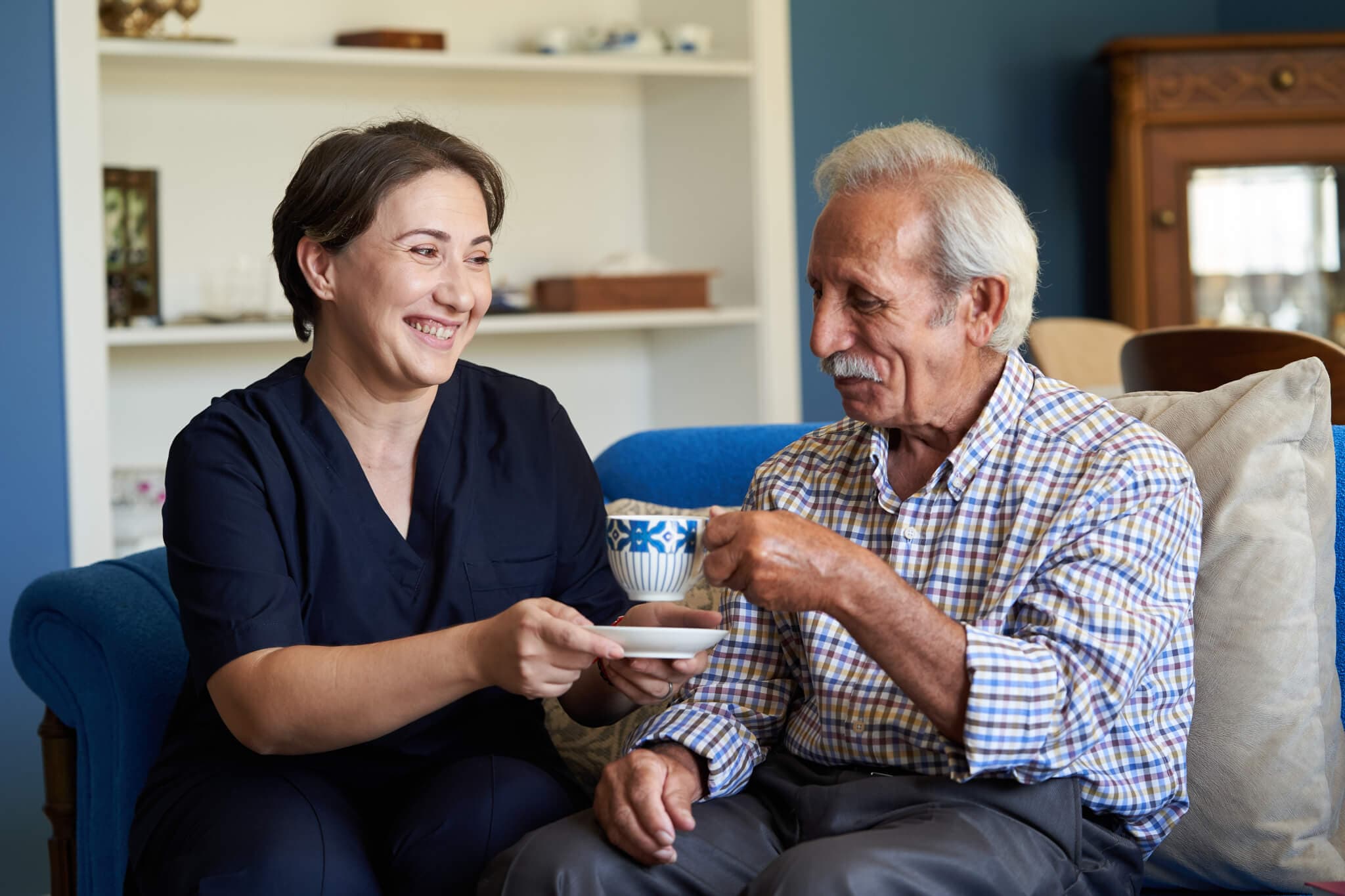 A caregiver smiles while handing a teacup to an elderly man sitting on a couch in a cozy living room. - Home Instead