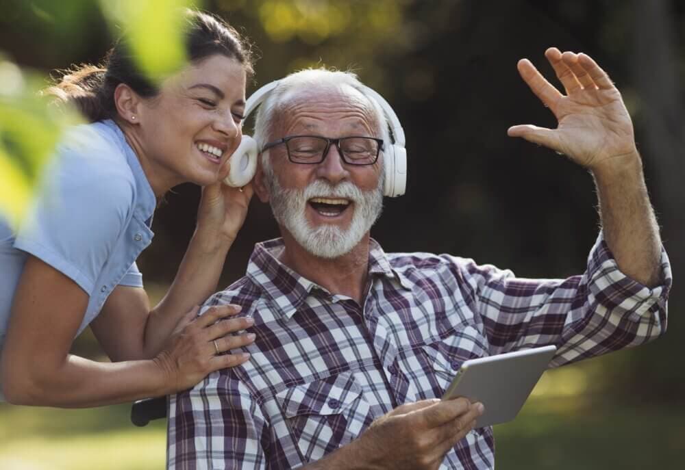 Elderly man wearing headphones and holding a tablet, laughing with a woman beside him in a outdoor setting. - Home Instead