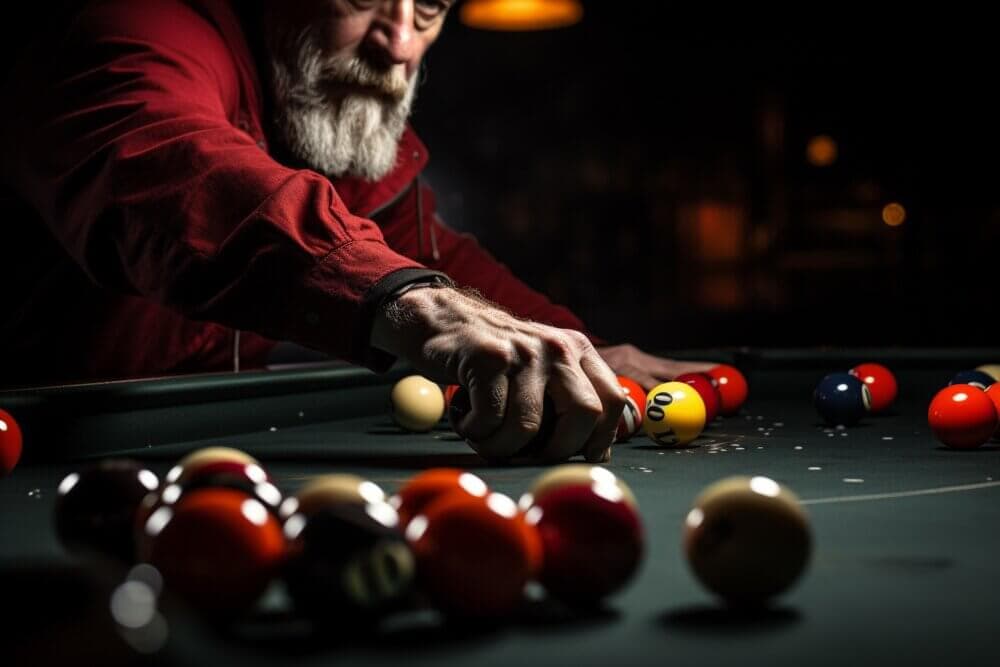 A bearded man in a red jacket playing pool, reaching to place the white cue ball on a dimly lit table. - Home Instead