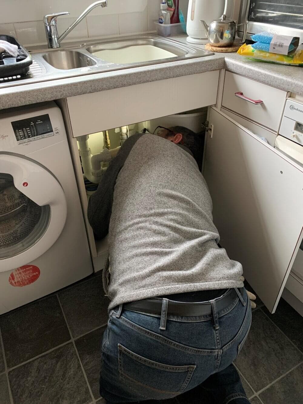 Person fixing plumbing under a kitchen sink, with tools and cleaning supplies around. - Home Instead