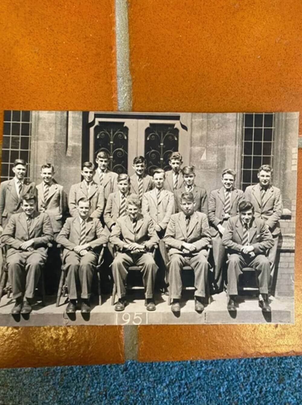 A black-and-white photograph of a group of 15 men in suits from 1950, posed sitting and standing outside a building. - Home Instead