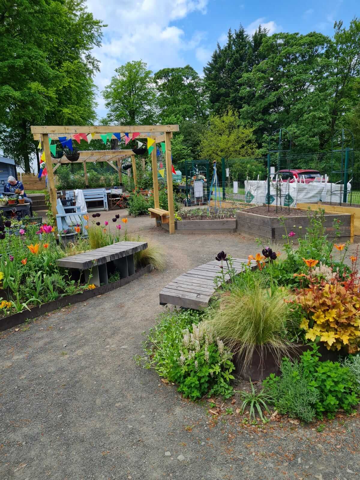 Colourful garden with benches, flowers, a swing, and a wooden pergola decorated with flags, surrounded by lush green trees. - Home Instead Renfrewshire and Barrhead community garden plot at Paisley's Barshaw Park.
