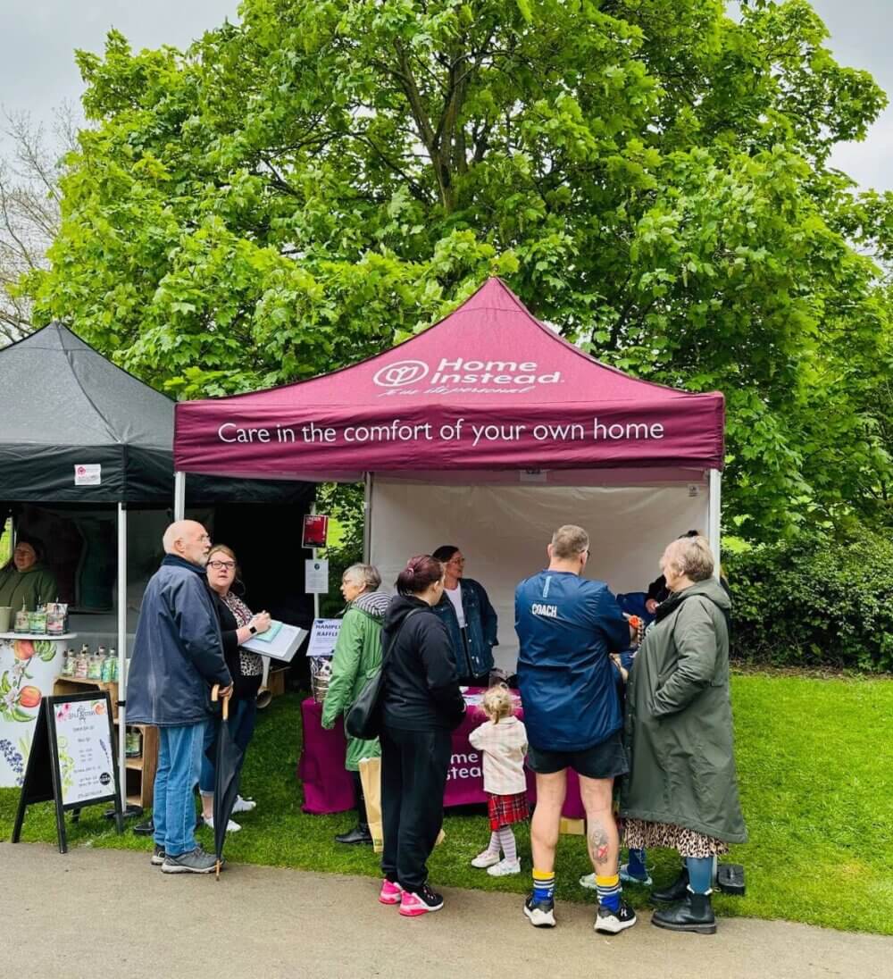 People gather near a Home Instead tent at an outdoor event; the tent offers home care information. - Home Instead