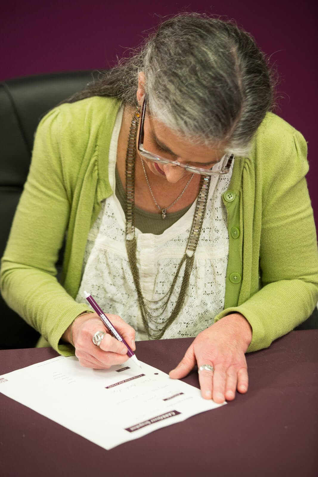 An elderly woman with gray hair signs a paper at a desk, wearing glasses, a green cardigan, and white blouse. - Home Instead