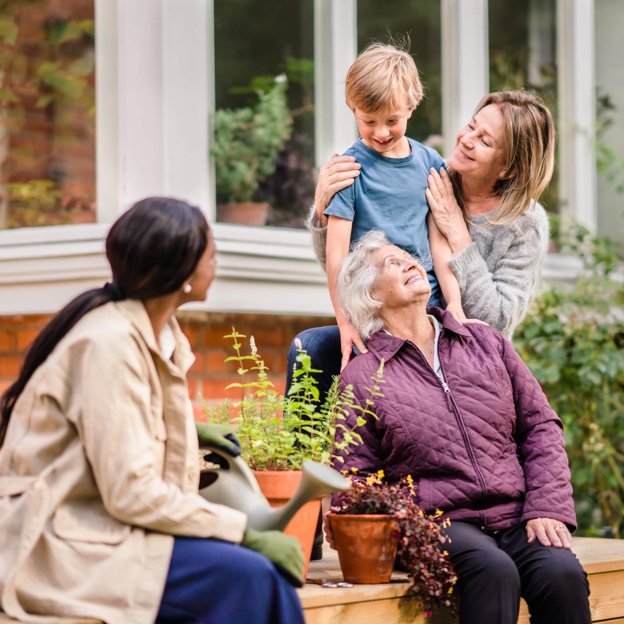 A young boy, his mom, and his grandmother sharing a happy moment while another woman waters plants nearby. - Home Instead