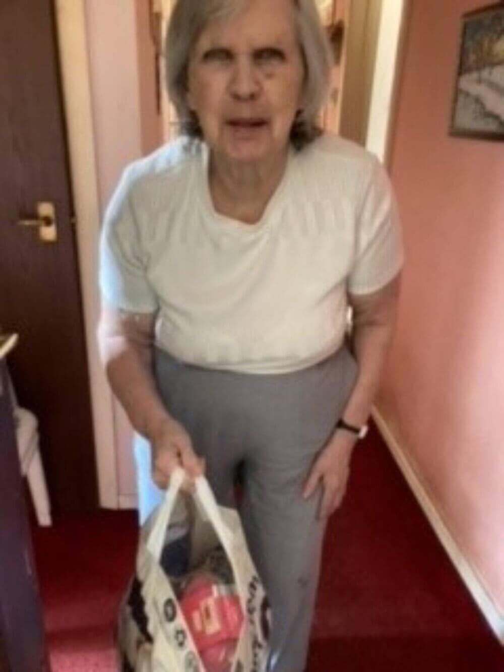 Person with gray hair in a white shirt and gray pants holding a grocery bag, standing in a hallway. - Home Instead