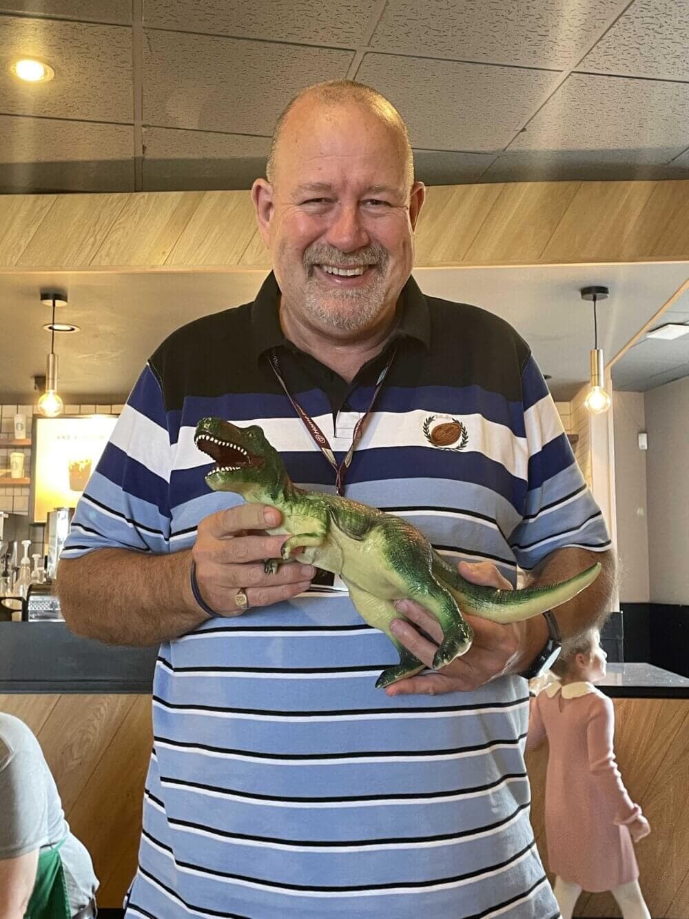 A smiling man holds a toy dinosaur in a casual cafe setting, with a blurred figure in the background. - Home Instead
