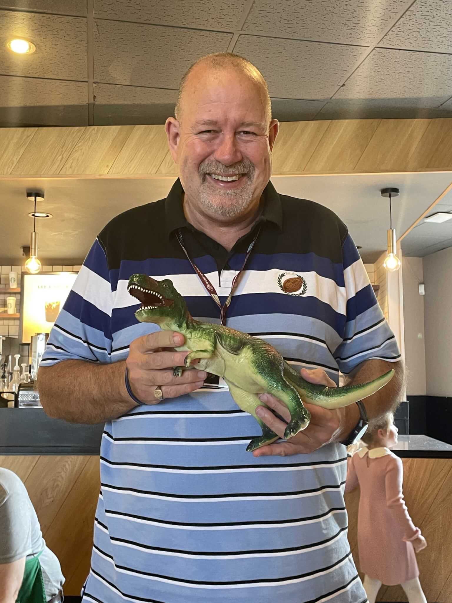 A smiling man holds a toy dinosaur in a casual cafe setting, with a blurred figure in the background. - Home Instead