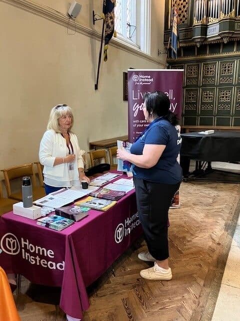 Two women talking at a Home Instead booth with brochures on a table inside a hall. - Home Instead