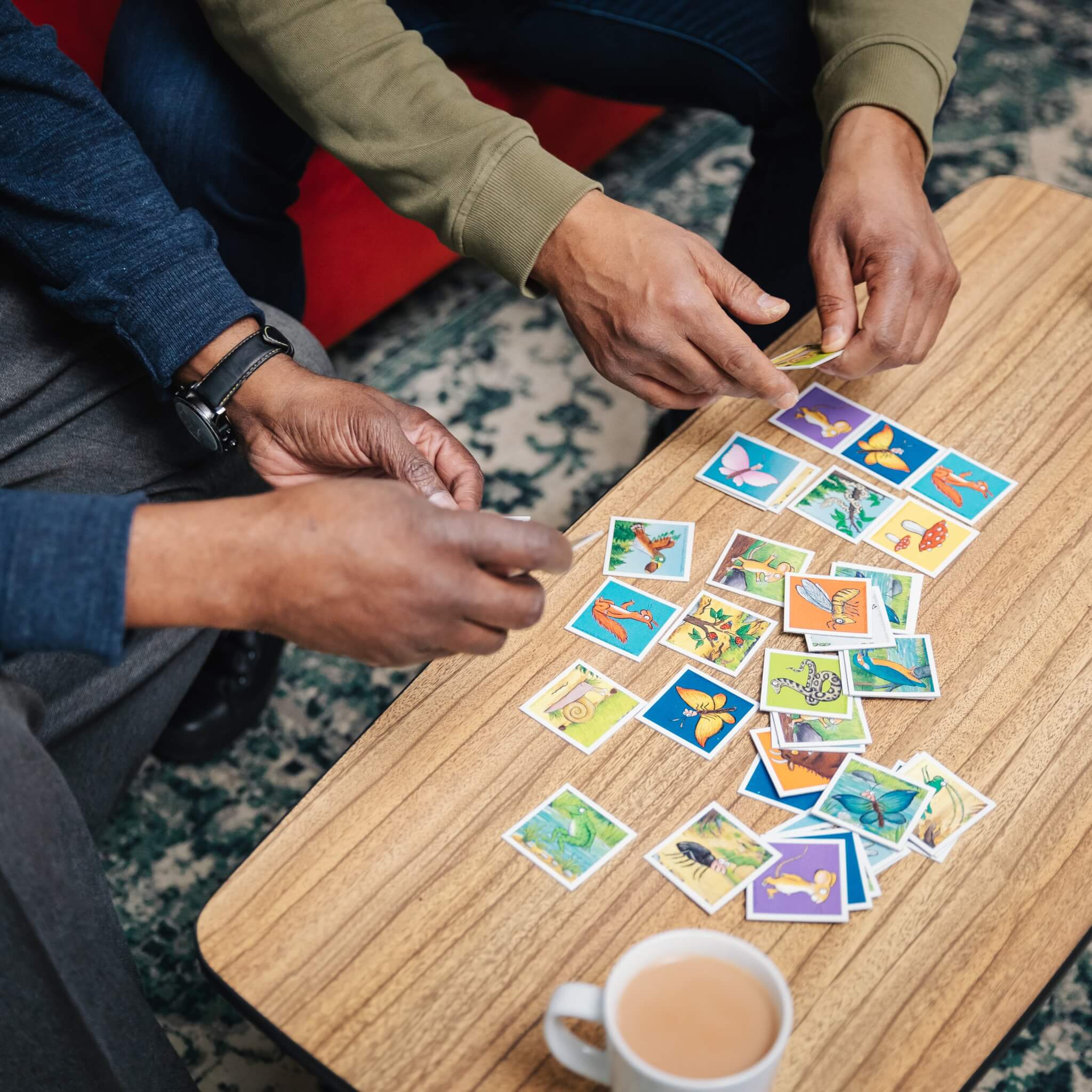 Two people sorting colorful animal cards on a table, with a cup of tea placed nearby. - Home Instead