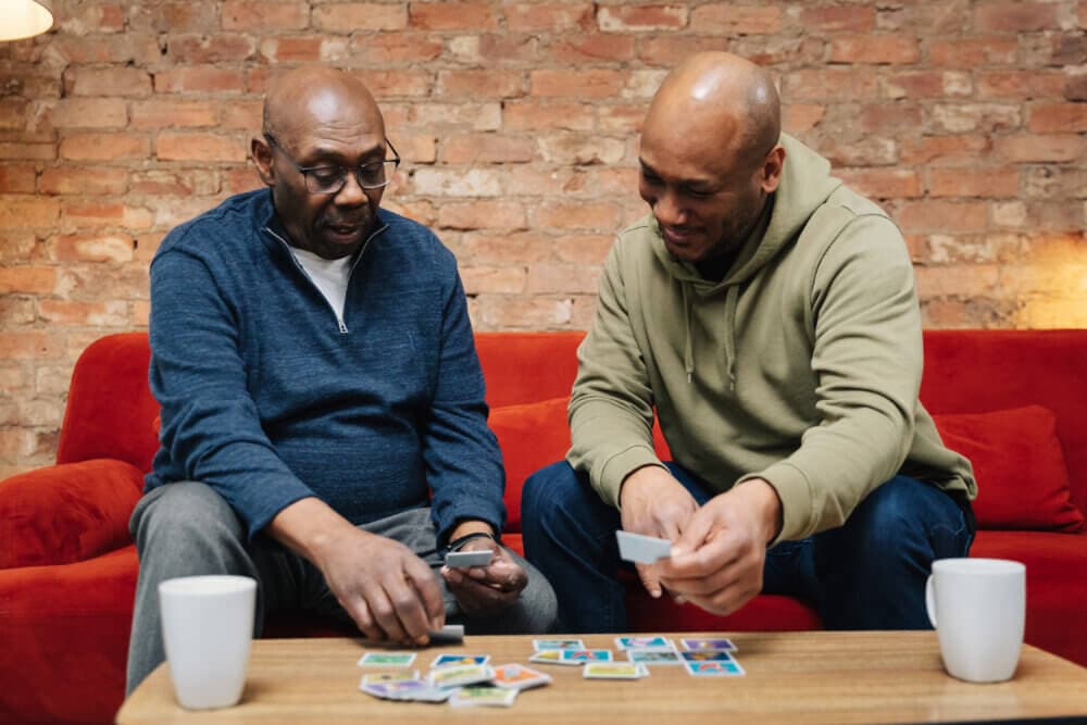 Two men sit on a red sofa, playing a card game on a coffee table, with two white mugs in front of them. - Home Instead