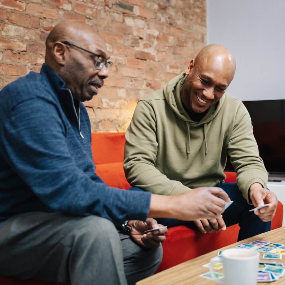 Two men sitting on a couch playing a card game, smiling and enjoying their time together. - Home Instead
