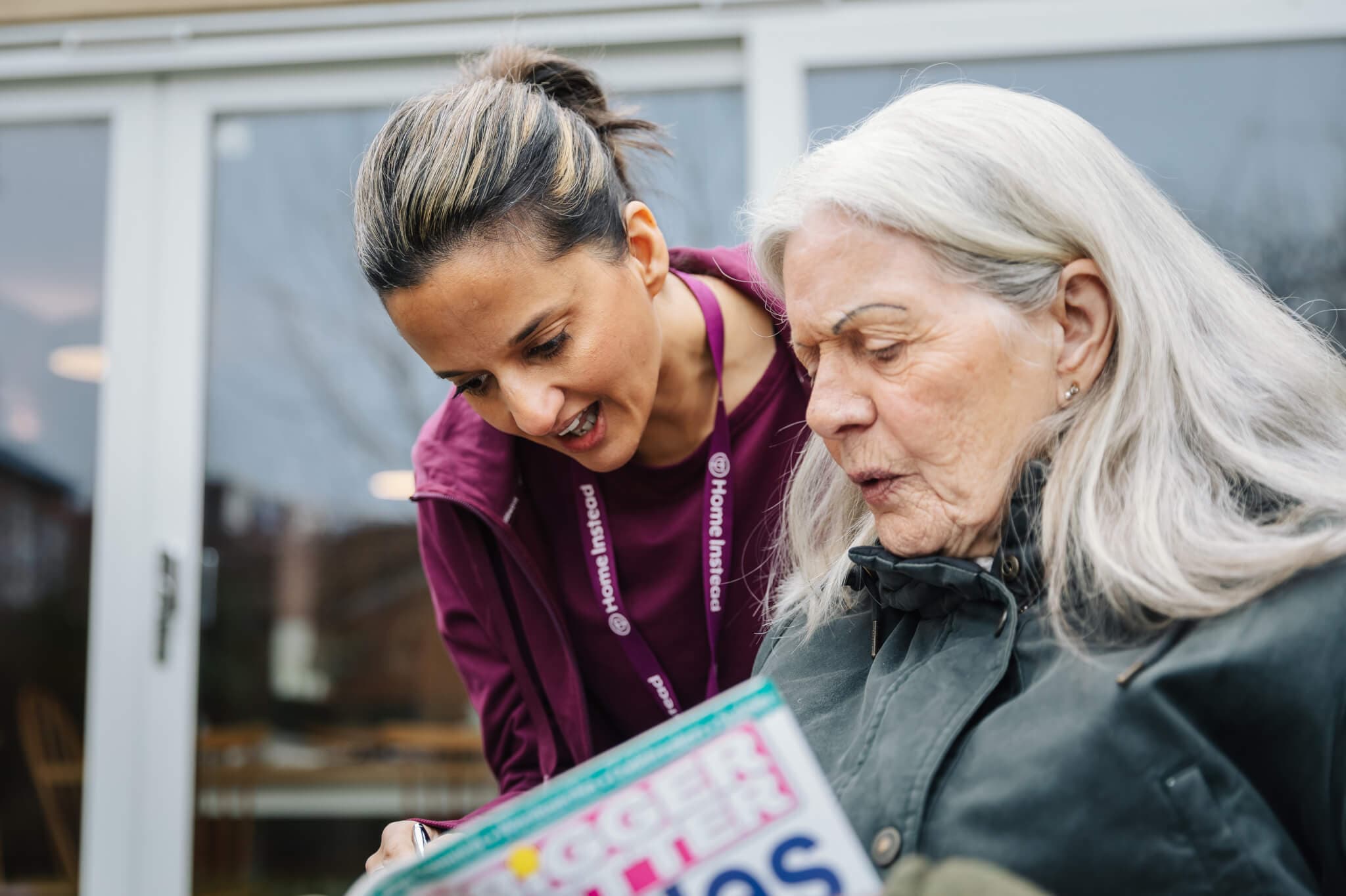 A caregiver smiles and looks at a puzzle book with an elderly woman outdoors in front of a building. - Home Instead