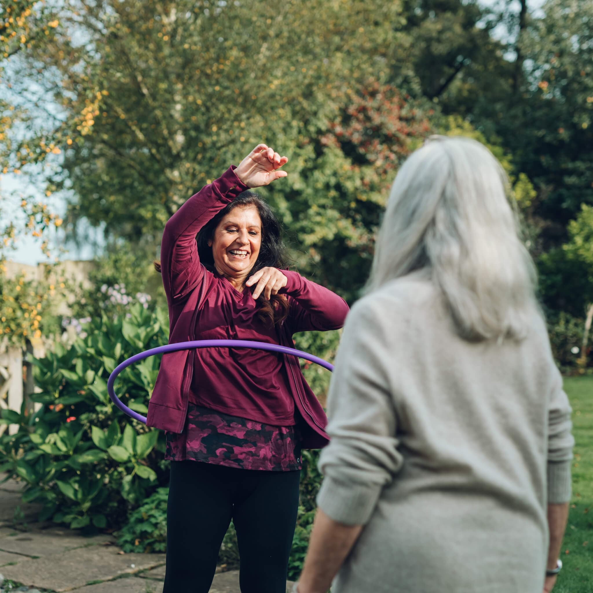 Two women outdoors, one hula hooping and smiling, while the other watches. Trees and foliage are in the background. - Home Instead