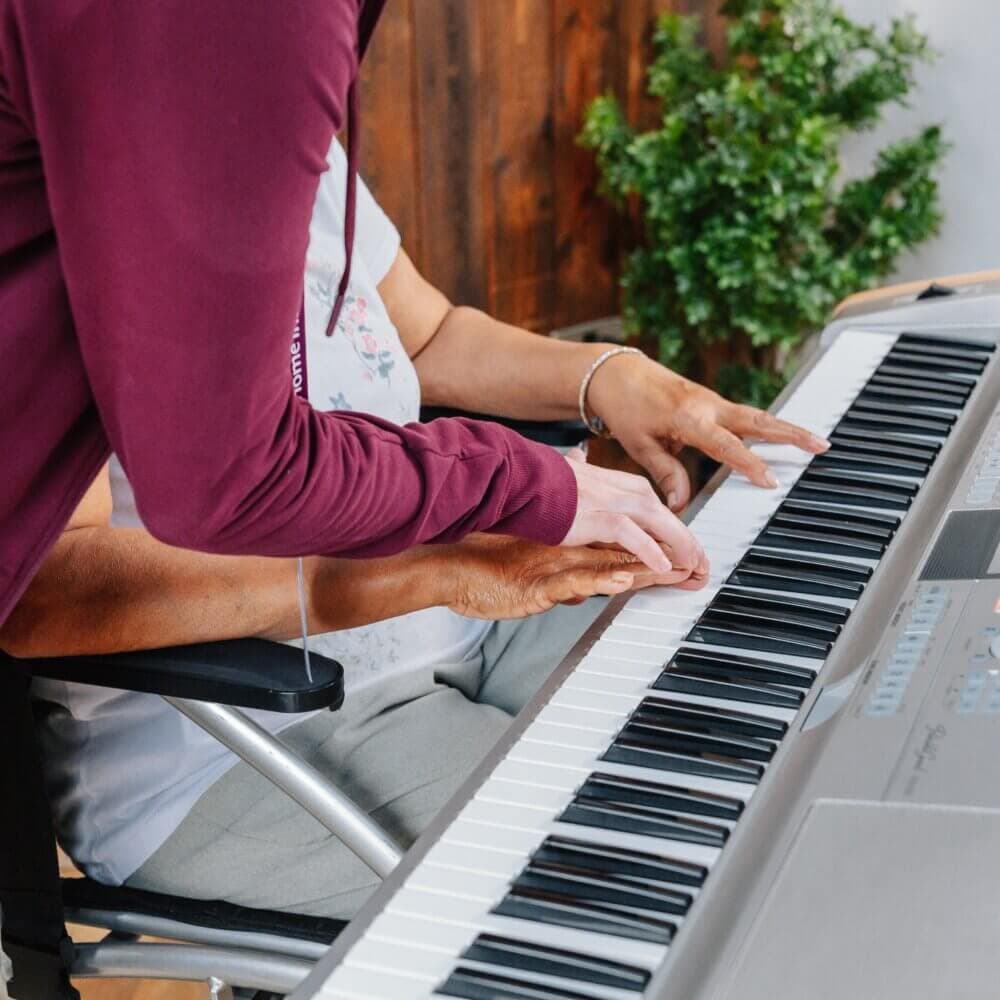 A person assists another person in playing an electric keyboard, with hands placed together on the keys. - Home Instead