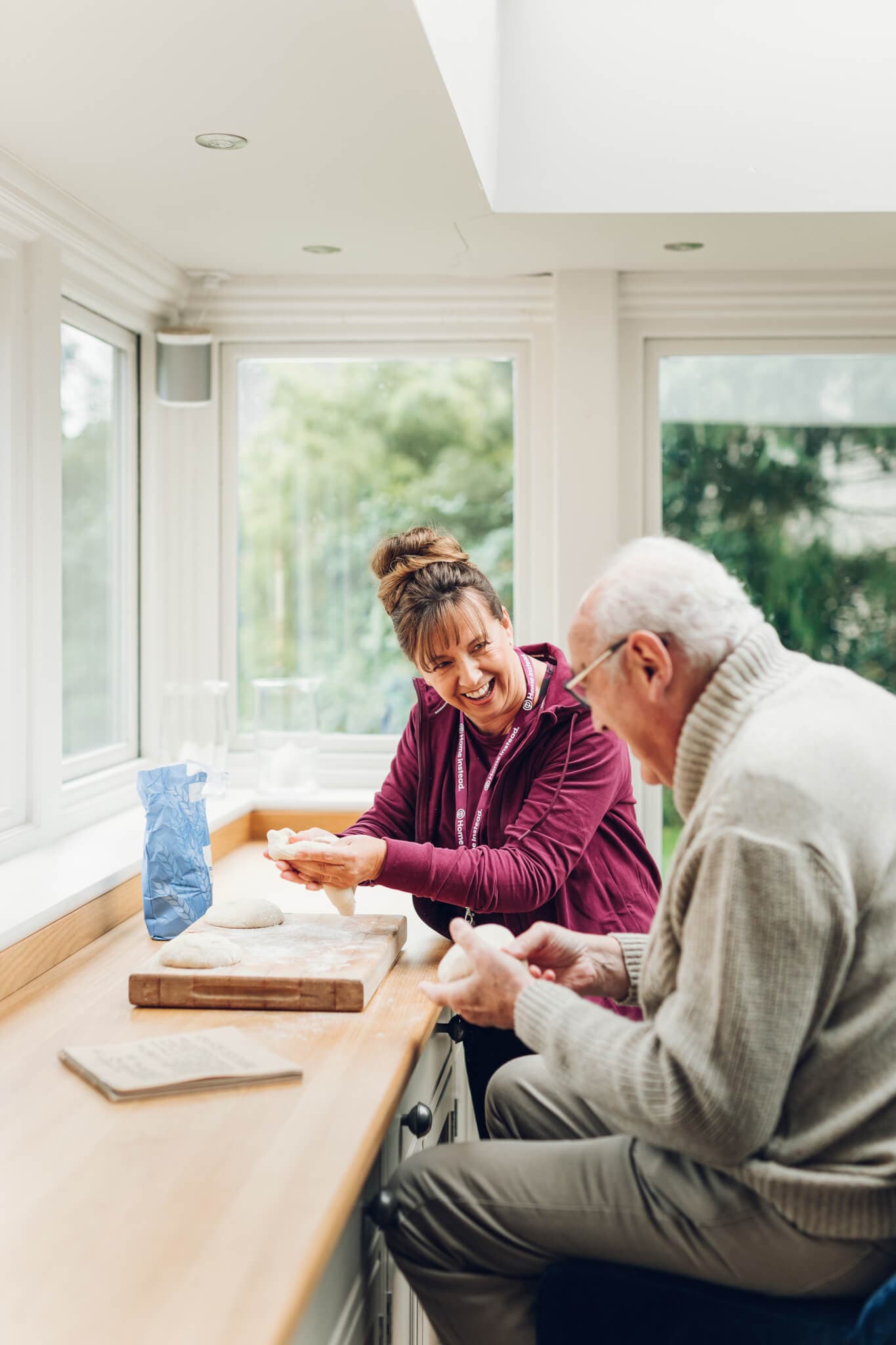 An elderly man and a woman smile and knead dough together on a kitchen counter by large windows. - Home Instead