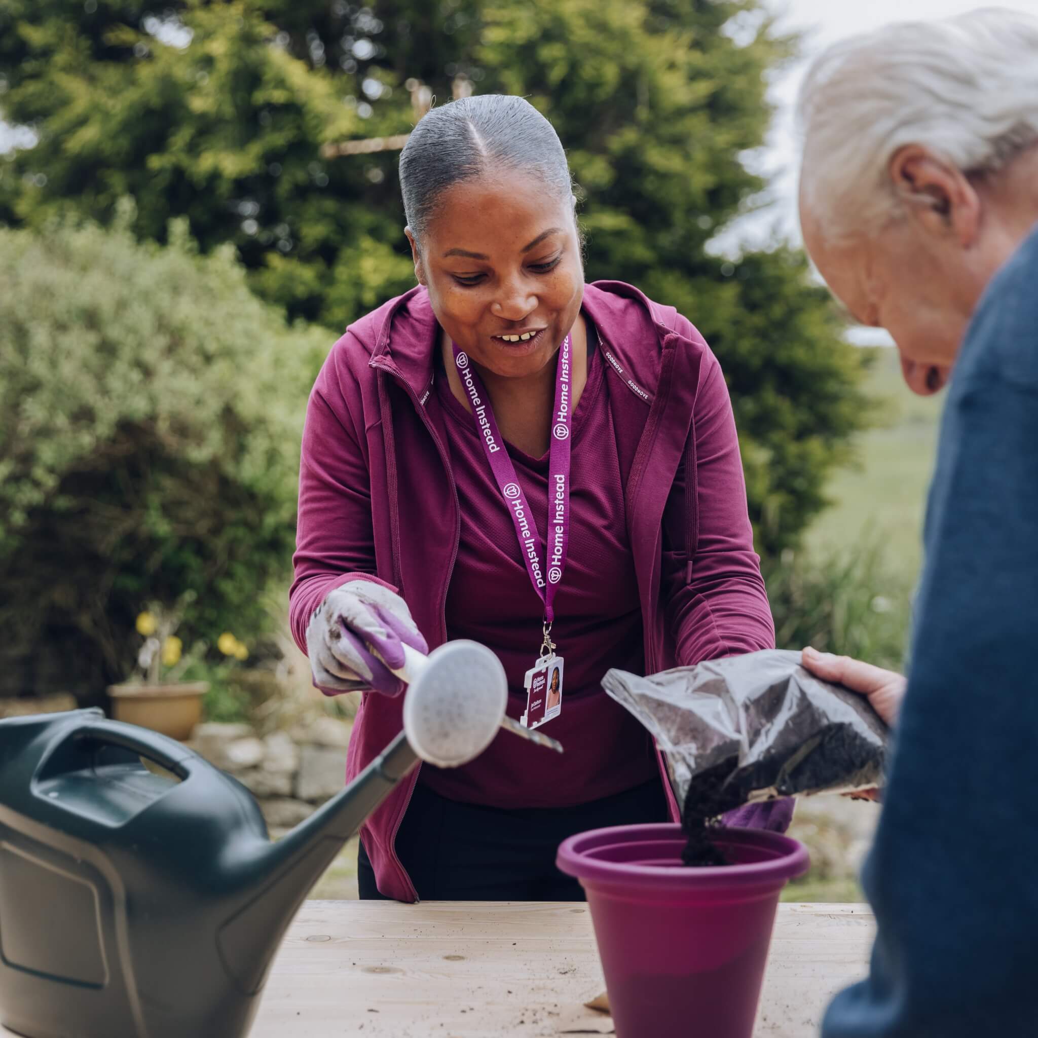 A woman helps an elderly man with gardening, pouring soil into a pot next to a watering can outdoors. - Home Instead