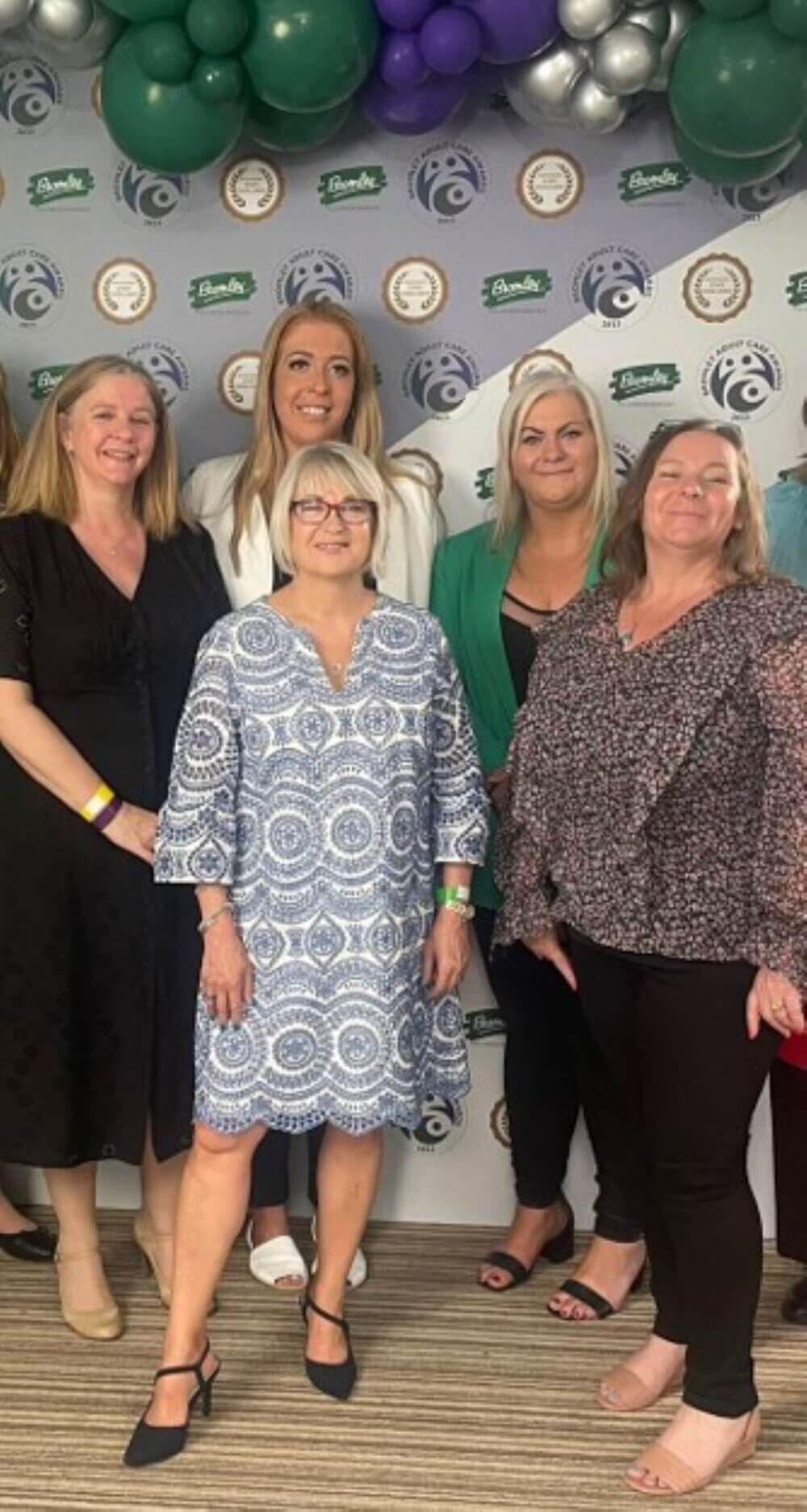 A group of five women smiling and posing together in front of a decorative backdrop with balloons. - Home Instead