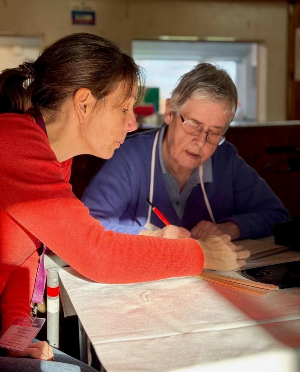 A woman in a red sweater helps an elderly woman with writing or drawing at a table. - Home Instead