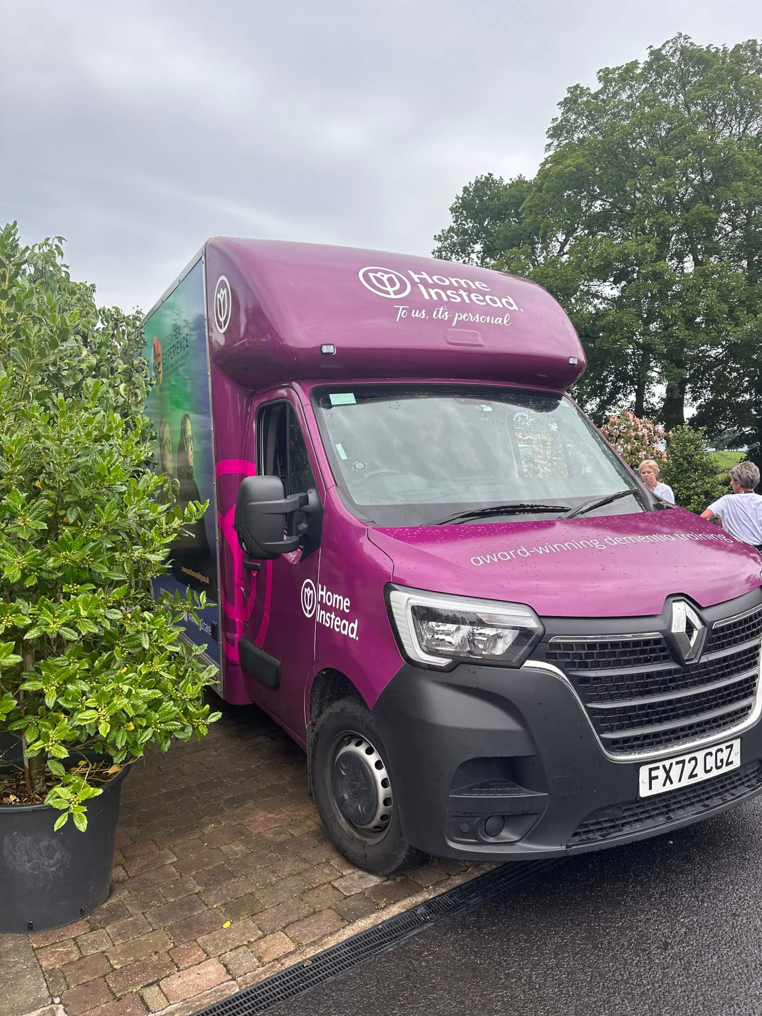 A purple Home Instead delivery van is parked on a driveway, surrounded by greenery and trees in the background. - Home Instead