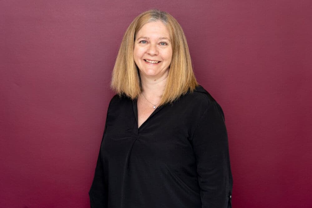 A woman with shoulder-length blonde hair smiles while standing in front of a red background, wearing a black blouse. - Home Instead
