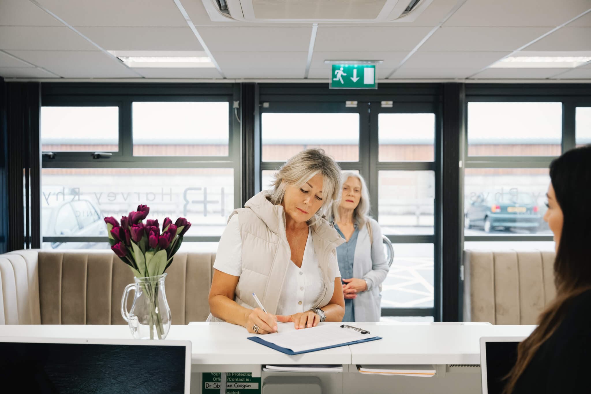 Two women at a reception desk. One woman is filling out a form while the other waits behind her. - Home Instead