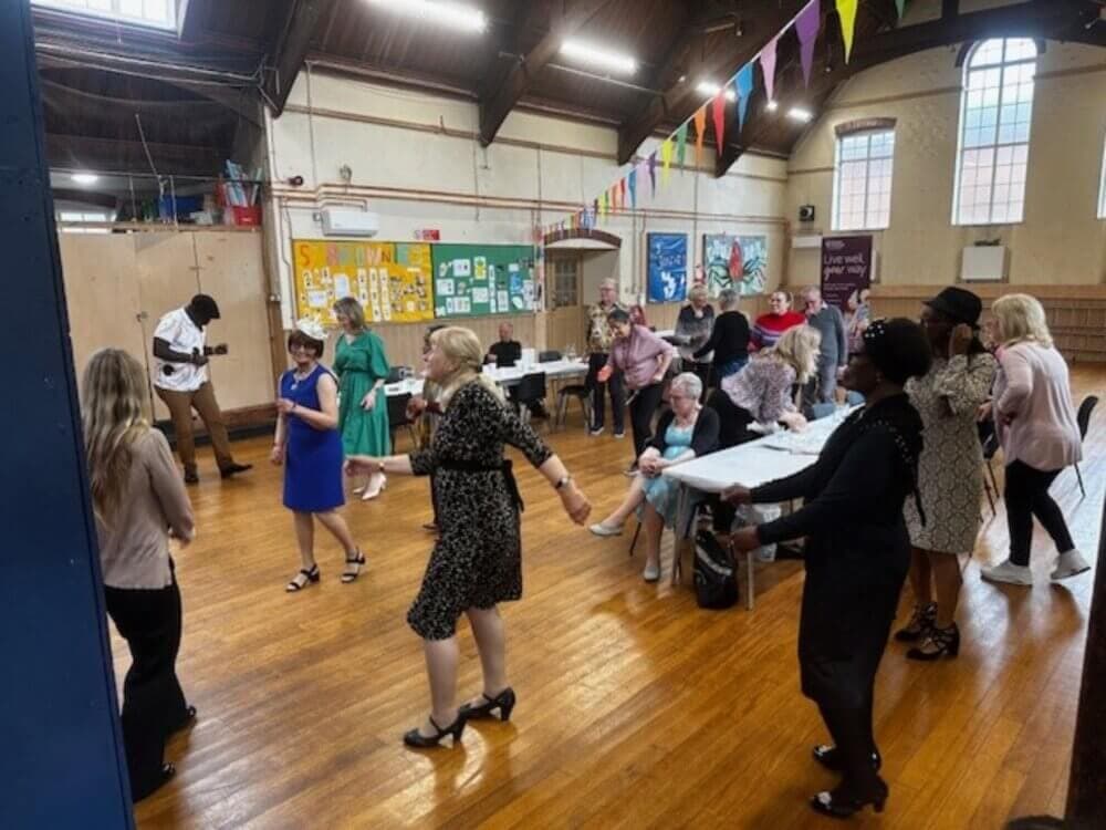 A group of people dancing and sitting at an indoor event with colorful flags hanging above and posters on the wall. - Home Instead
