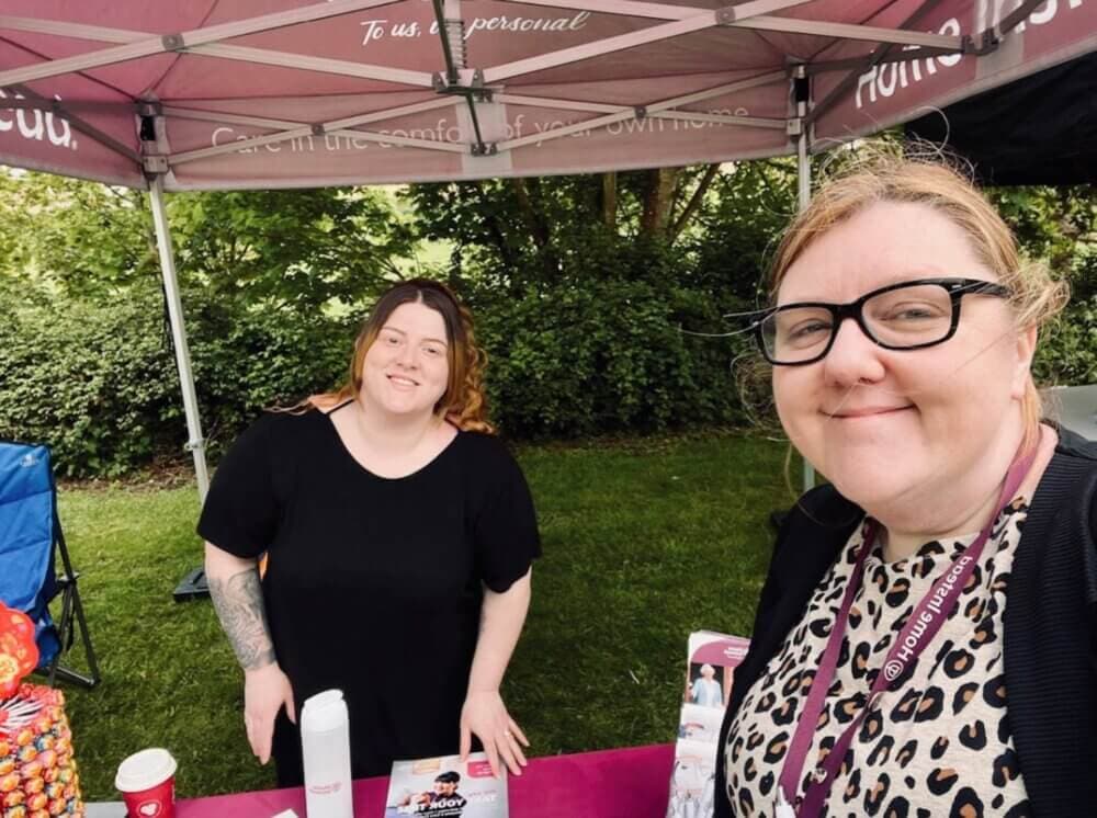 Two women smiling at a booth outdoors, one behind a table with brochures and candy, and the other taking a selfie. - Home Instead