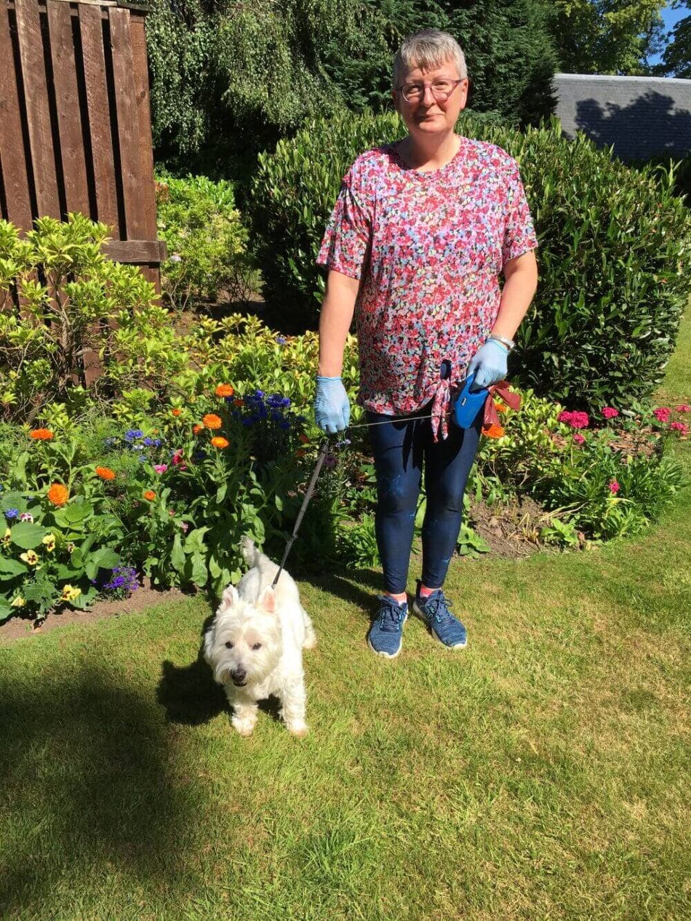 Person standing in a garden, holding the leash of a small white dog on a sunny day. - Home Instead