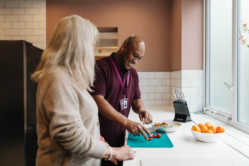 Two people sharing a moment in a kitchen, one slicing vegetables on a cutting board while the other watches and smiles. - Home Instead